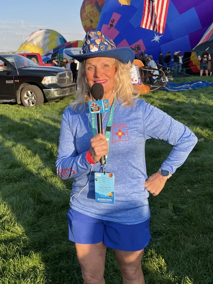balloon-fiesta-live-crewperson-with-mic A person in blue attire smiles, holding a microphone at the Balloon Fiesta field, surrounded by colorful balloons and other attendees.