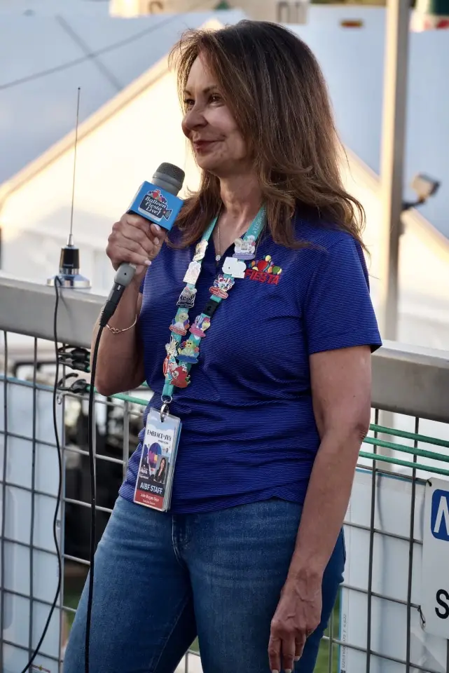 balloon-fiesta-live-crewperson-with-mic02 A person wearing an official blue balloon fiesta shirt speaks into a microphone with event badges. Background includes a metal railing and tent structure.