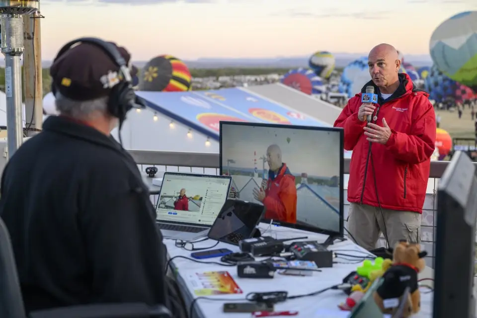 balloon-fiesta-live-crewperson-with-mic03 Two people are broadcasting from a booth at a hot air balloon festival. One person is holding a microphone, with balloons in the background.