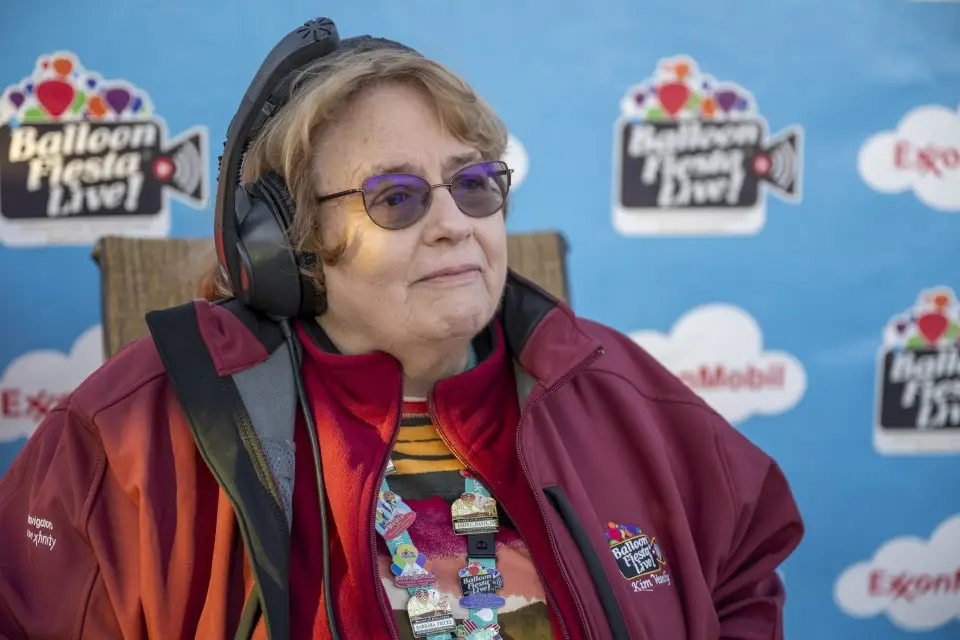balloon-fiesta-live-crewperson A person with headphones and jacket adorned with pins stands against a "Balloon Fiesta Live!" backdrop