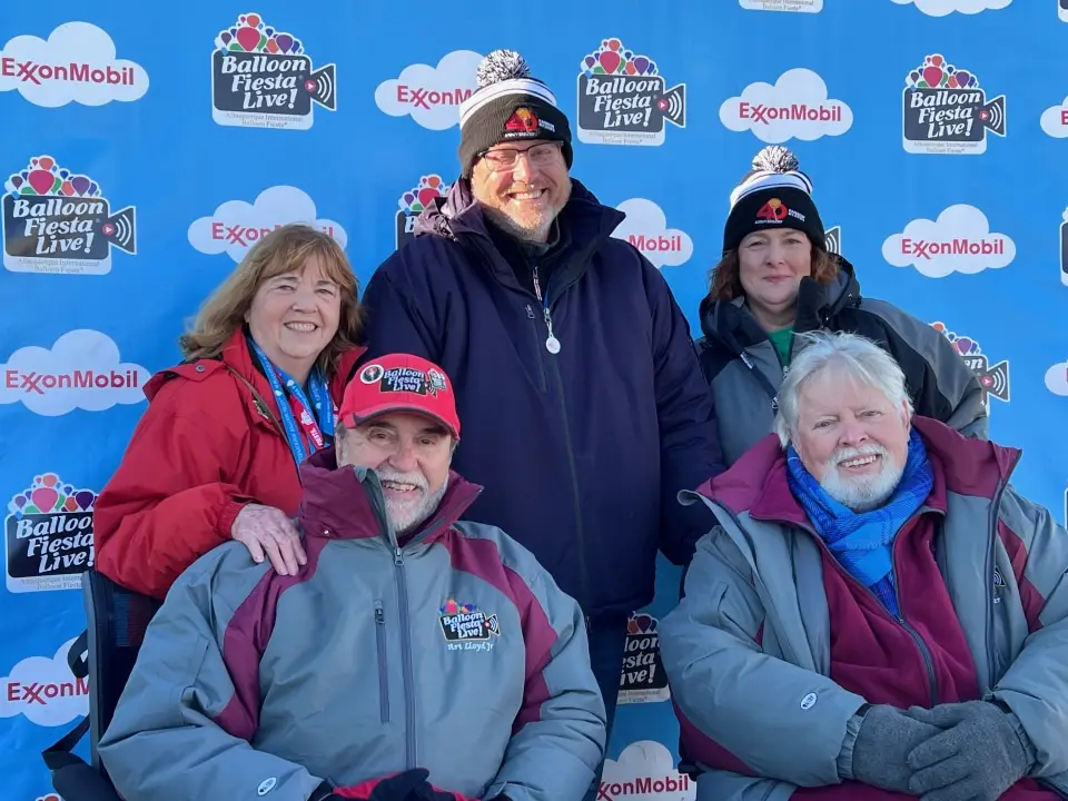 balloon-fiesta-live-group-photo Group of five people in winter clothing, smiling, posing in front of a "Balloon Fiesta Live!" backdrop with logos and a blue background.