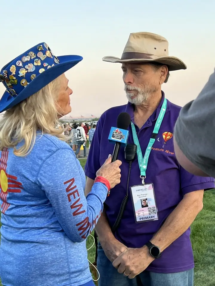 balloon-fiesta-live-interview Two people standing outdoors, person with microphone interviewing another person wearing a hat. Person's shirt and hat have colorful designs and text.