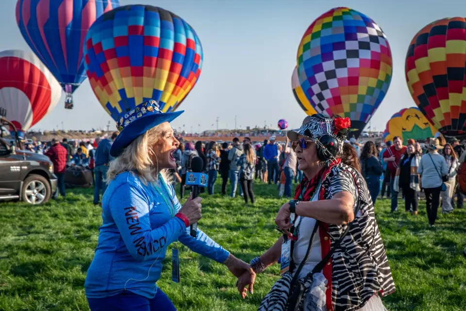 balloon-fiesta-live-interview02 Balloon Fiesta Live crewmembers talking into microphone near colorful hot air balloons on balloon fiesta grounds, with a lively crowd in the background on a sunny day.