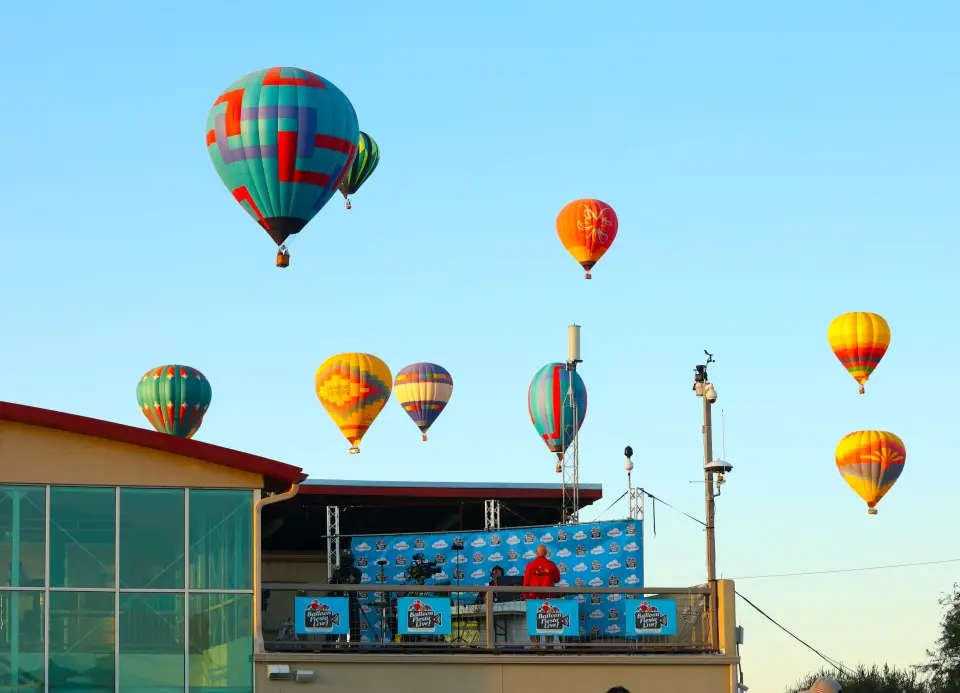 balloons-floating-by-studio A person overlooks colorful hot air balloons floating in clear skies at Albuquerque Balloon Fiesta, with equipment visible on a building rooftop.