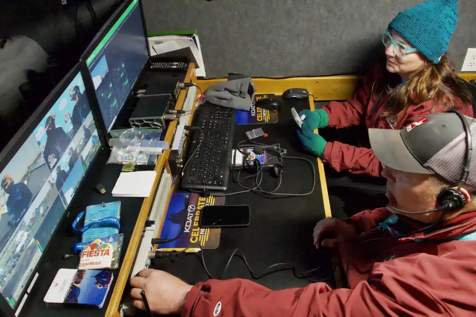 inside-the-production-truck Two people in a control room monitor screens, managing a live broadcast. Tech gadgets and event passes are scattered on the desk.