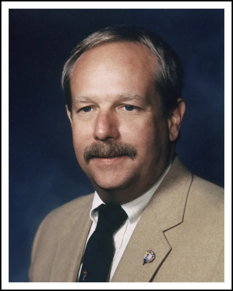 JohnC.DavisIV A person with a mustache is wearing a beige suit and tie, posed against a dark blue background in a formal portrait.